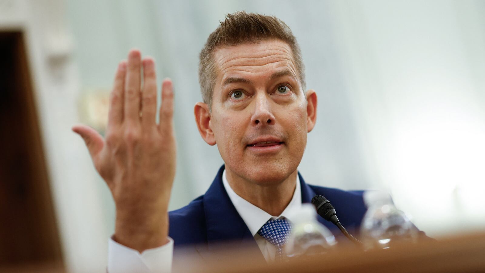 Sean Duffy sits at a table, speaking, gesticulating with one hand.