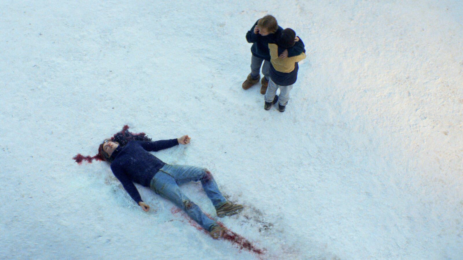 A dead body lies on the snow, blood pooled under his head, as a woman and child hug nearby.