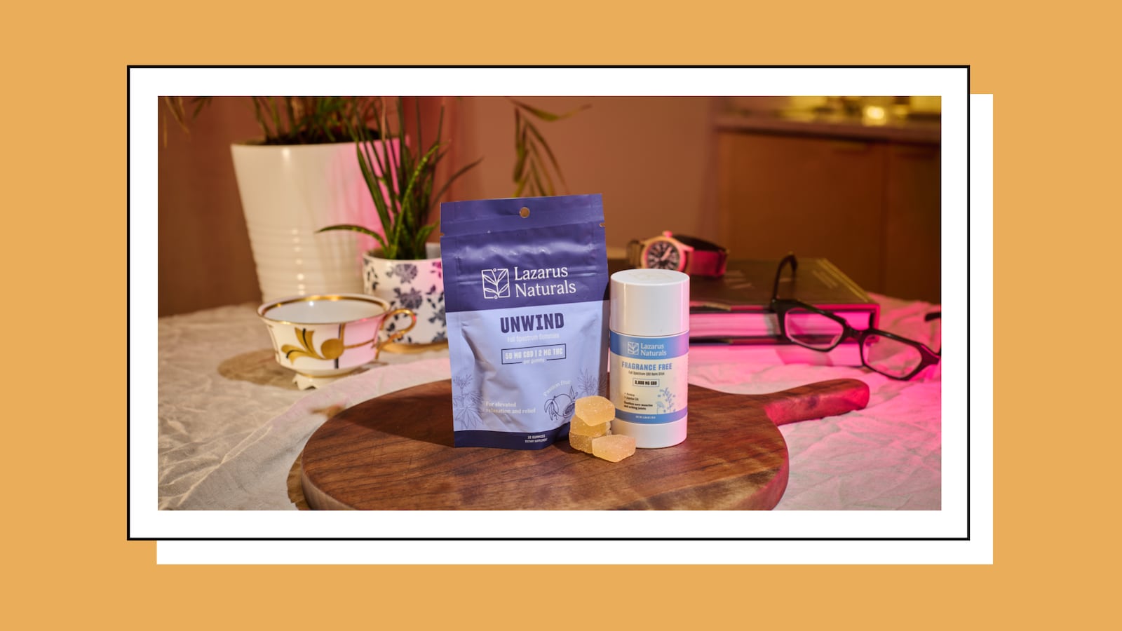 A product photo featuring Lazarus Naturals CBD items displayed on a wooden board. In the foreground is a blue resealable bag labeled “Unwind” and a white cylindrical stick labeled “Fragrance Free,” with three yellow gummy cubes placed in front. The background includes a teacup, potted plants, a watch, glasses, and books on a table with warm and pink lighting.