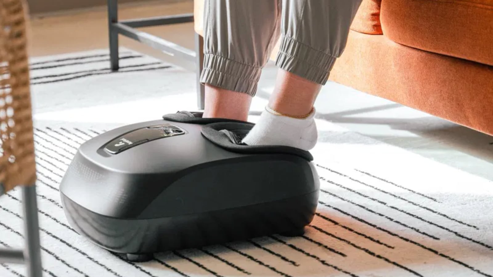 A person using the AuraHeal foot massager. They are sitting down on a couch and have both feet in the massager. They have grey pants and white socks on.