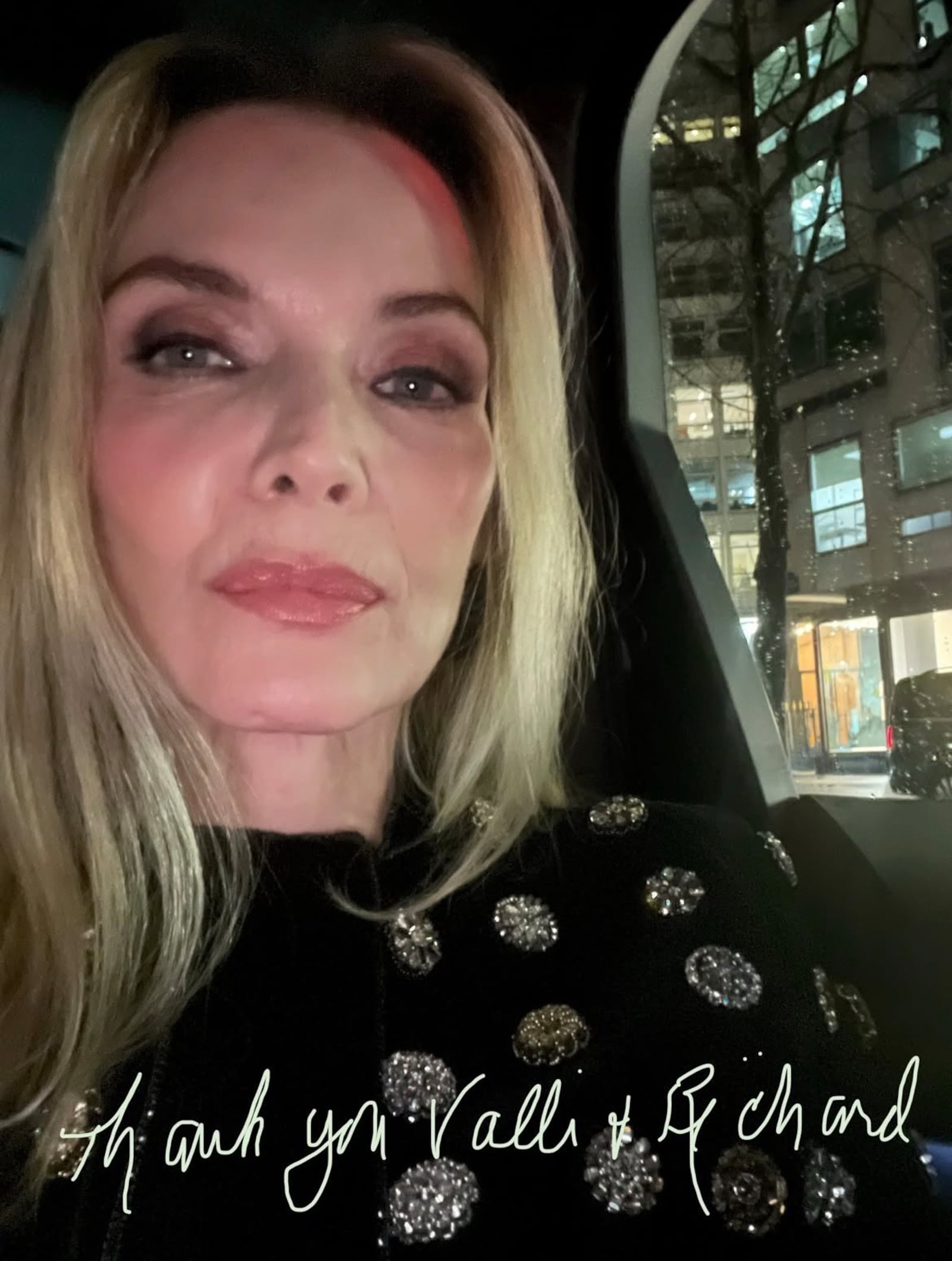 Michelle Pfeiffer wearing a dark smoky eye, lip gloss, and a sparkly top as she poses in the backseat of a car.