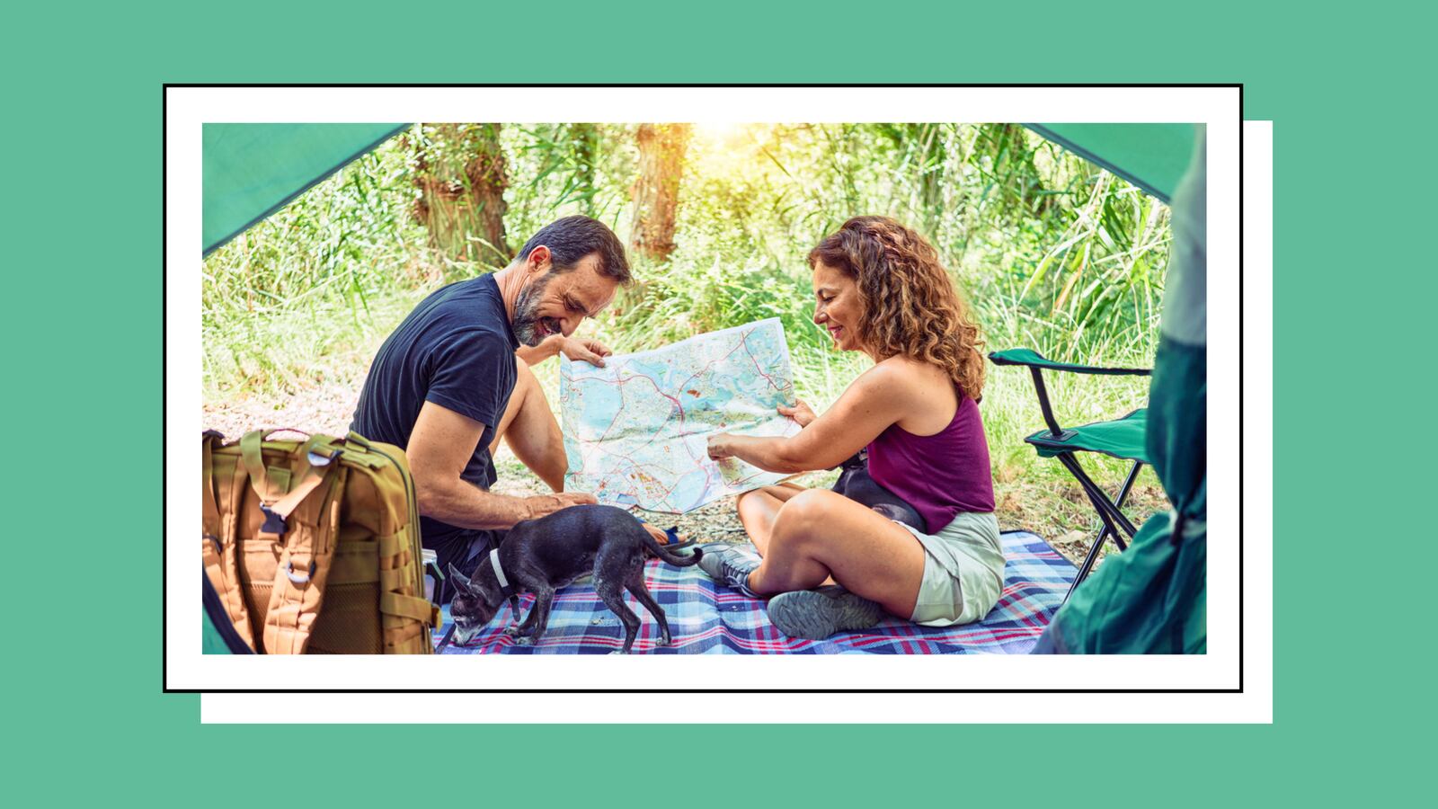 A middle-aged couple looks at a map as they sit outside on a plaid blanket in front of their tent. A cute black dog moves across the frame.