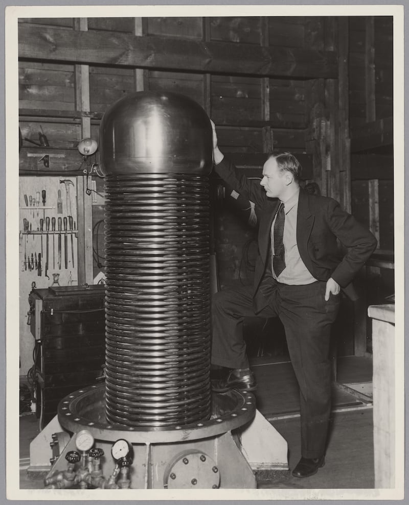Black and white picture of a man in a dark suit standing behind a large metal object which appears to be like a generator