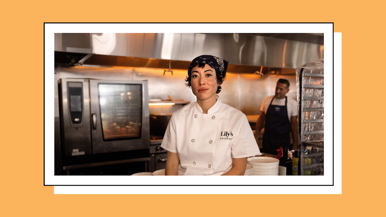 A chef wearing a white double-breasted coat embroidered with “Lily’s Catering” and a navy bandana stands in a commercial kitchen, facing the camera. Stainless steel ovens and warm overhead lights glow behind her, while another staff member works in the background near metal racks and kitchen equipment.
