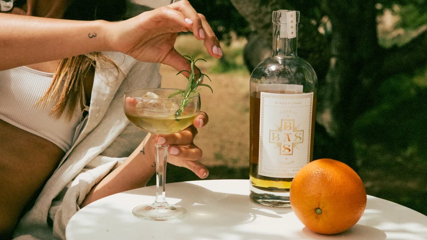 Bottle of Basbas herbal liqueur next to a cocktail and an orange on a table.