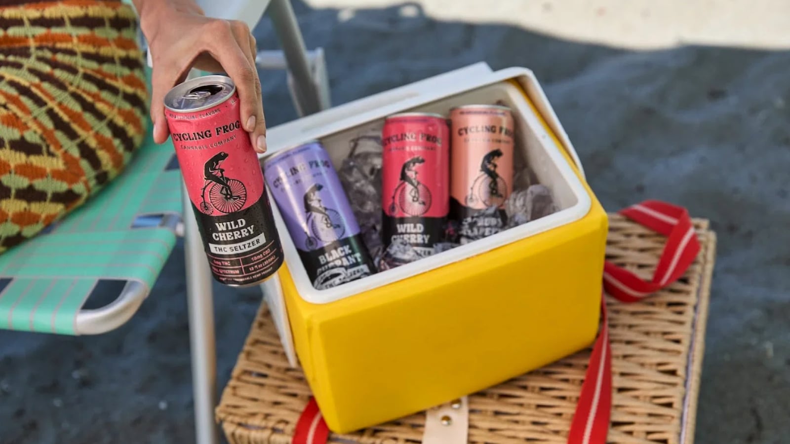 A hand reaches into a yellow cooler filled with ice and Cycling Frog Cannabis Company THC seltzer cans in multiple flavors, including Wild Cherry and Black Currant. The cooler sits on a wicker basket outdoors, suggesting a warm-weather gathering or beach setting