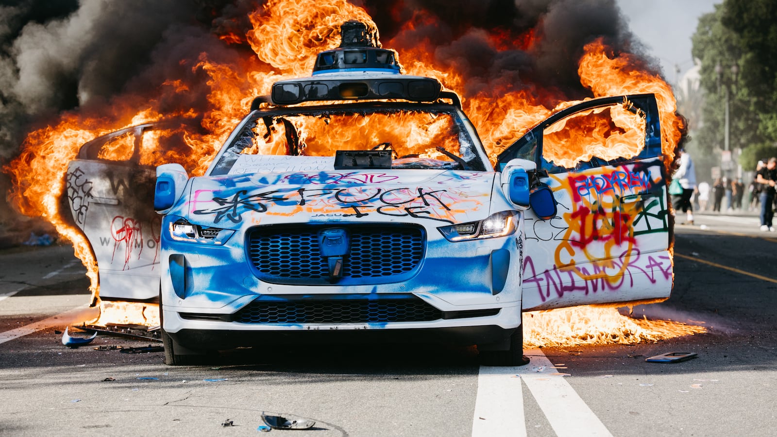 A Waymo car burns in the street as smoke billows during protests in Los Angeles, California, on June 8, 2025. Following a series of aggressive federal immigration operations in Los Angeles, tensions escalated when Immigration and Customs Enforcement (ICE) agents conducted a high-profile raid on a Home Depot location. The raid sparked widespread protests across the city, where demonstrators decried the targeting of immigrant communities and the separation of families. Clashes soon broke out between protesters and federal agents, prompting U.S. President Donald Trump to deploy 2,000 National Guard troops to the city, a move harshly criticized by California officials, including the governor, as "purposefully inflammatory." (Photo by David Pashaee / Middle East Images via AFP) (Photo by DAVID PASHAEE/Middle East Images/AFP via Getty Images)