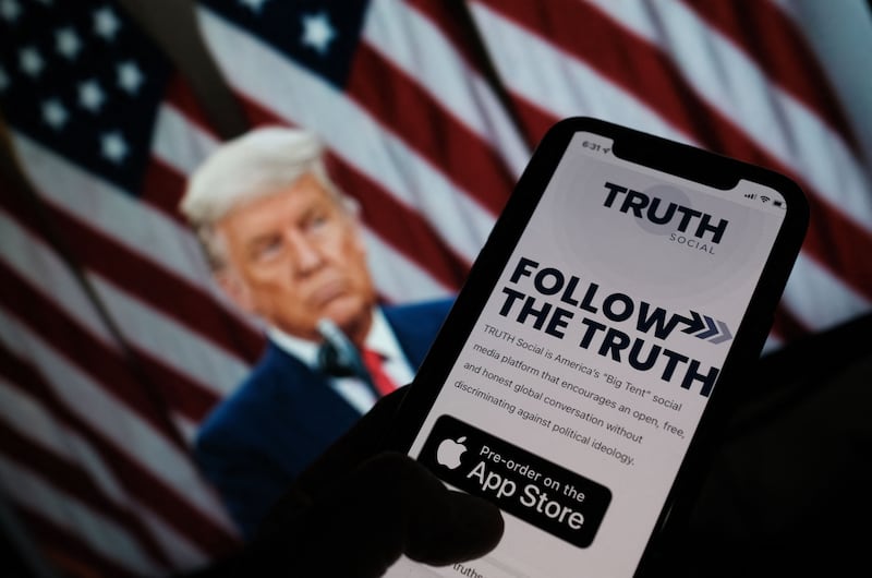 This illustration photo shows a person checking the app store on a smartphone for "Truth Social", with a photo of Donald Trump on a computer screen in the background.