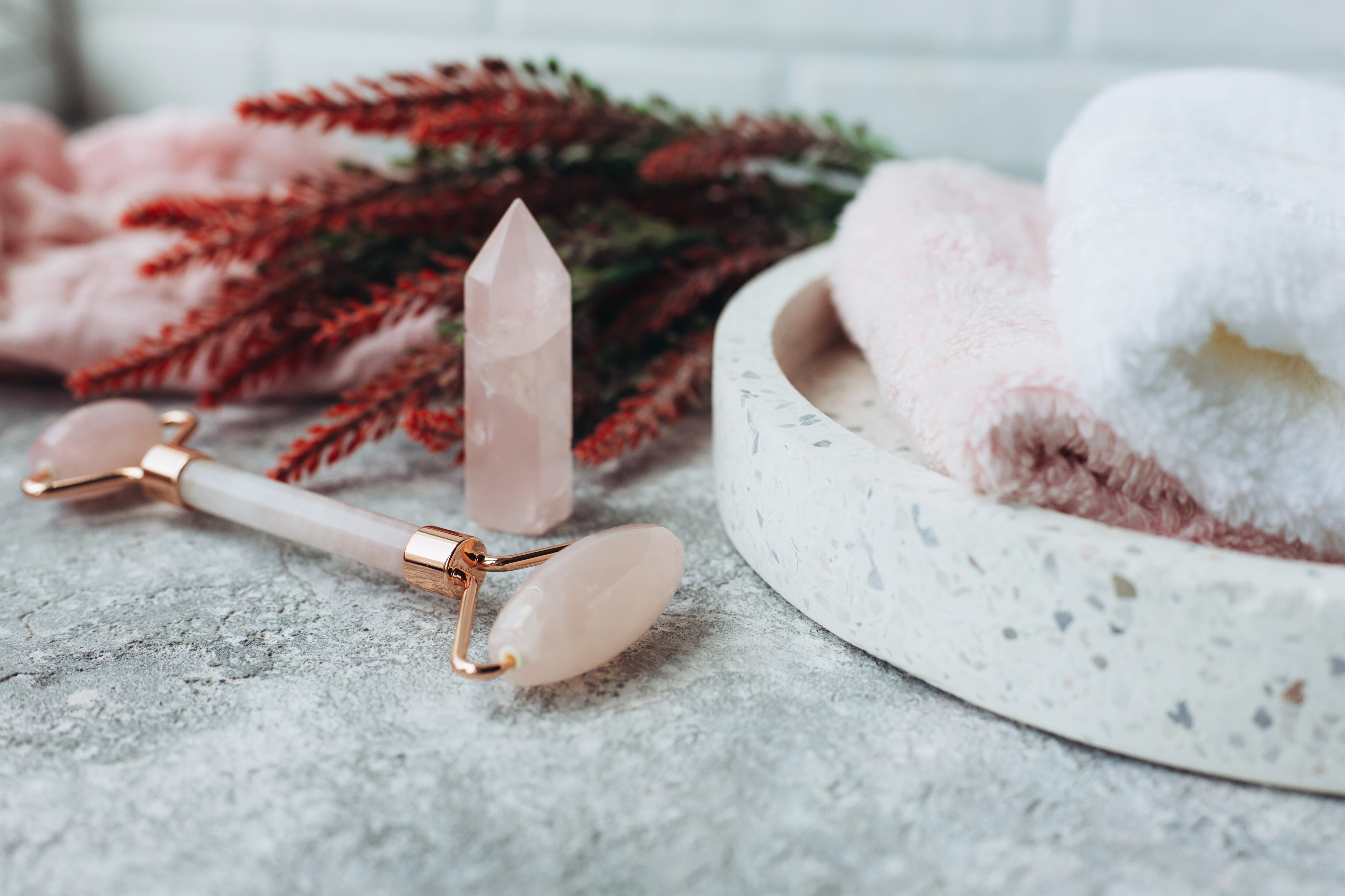 Rose gua sha roller and quartz near tray with towels on a light surface with artificial twigs.