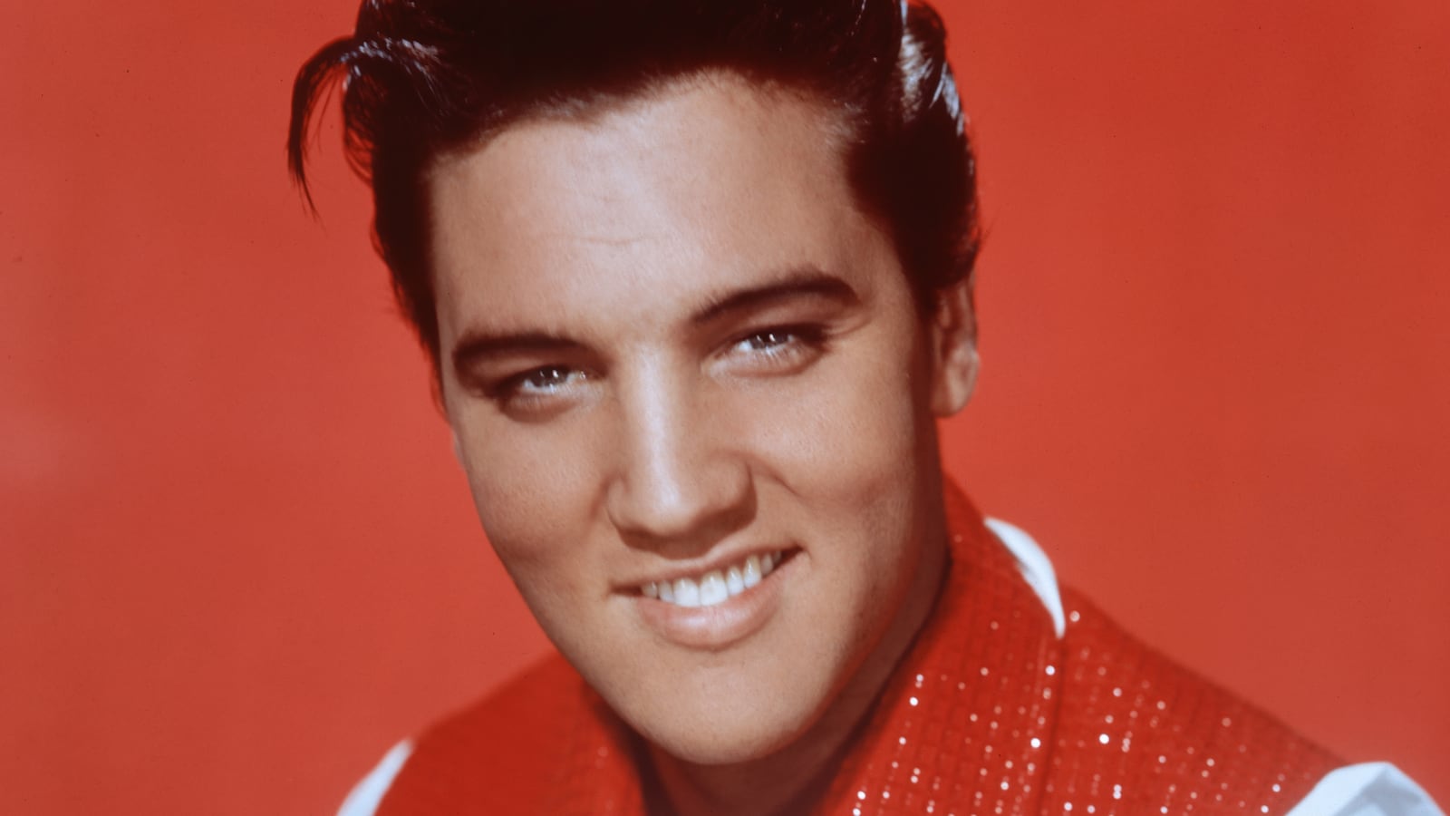 Elvis Presley smiles in a portrait with a red waistcoat and background
