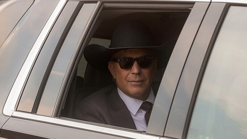 Kevin Costner wears sunglasses and a cowboy hat in a car with a rolled down window in a still from "Yellowstone"