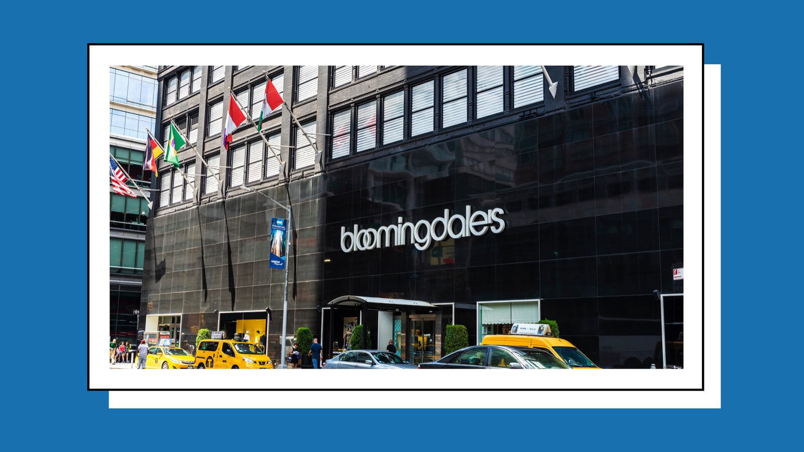 Front view of Bloomingdale's store exterior on a sunny day, featuring its iconic logo on a black facade. Several flags are displayed above, and yellow taxis are parked outside, with people walking by.