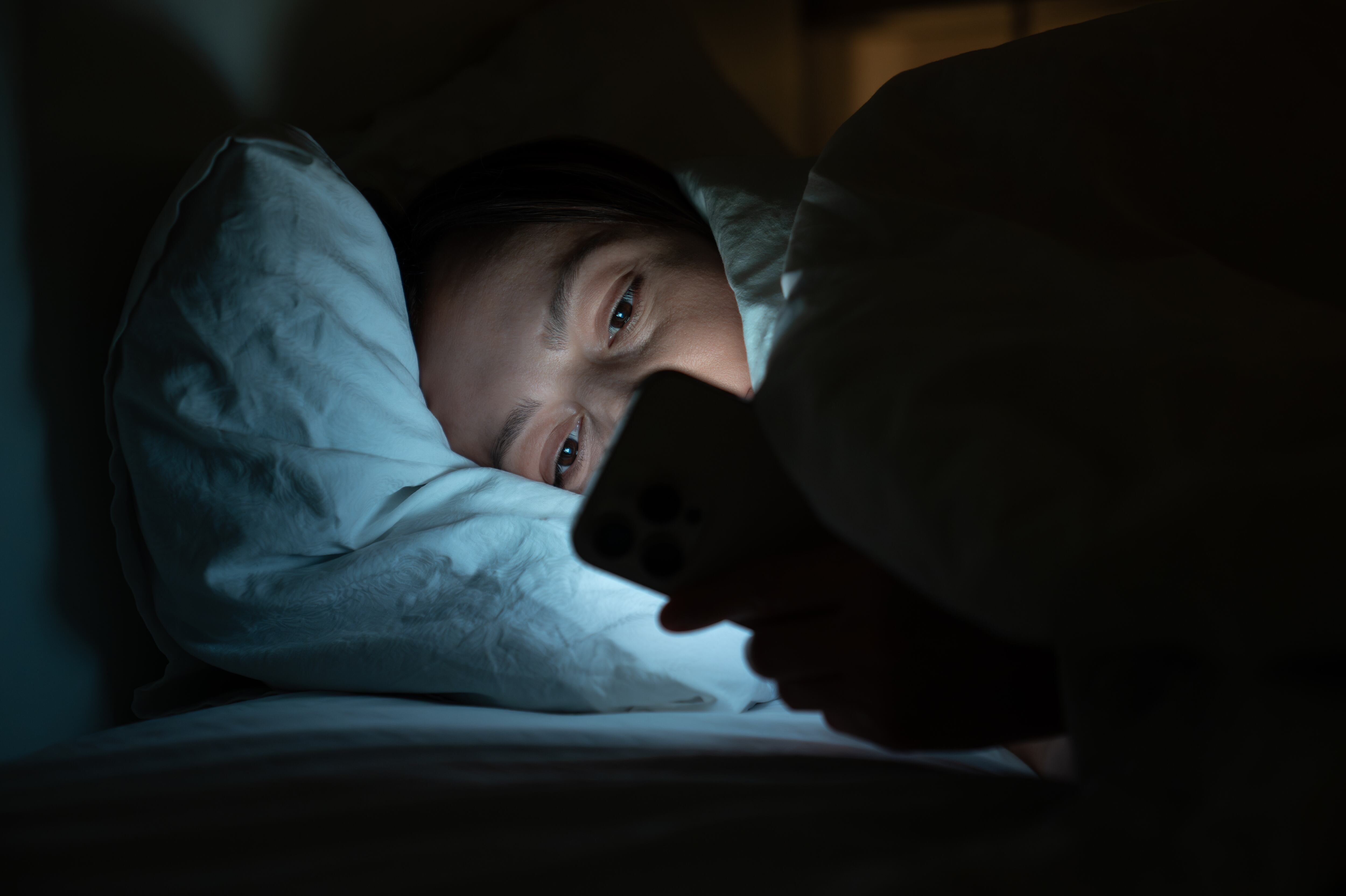 Sleepless woman having sleep deprivation lying in bed, looking at illuminated mobile phone screen. Unhappy female with insomnia surfing in internet, hanging in social networks, reading news, messaging