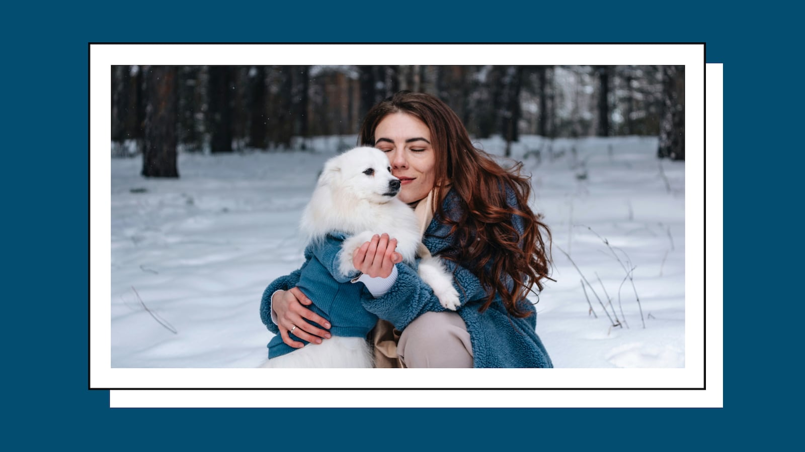 A woman with long brunette hair hugging her dog in the snow. They are both wearing matching blue jackets. In the background, you can see snow on the ground and a forest.