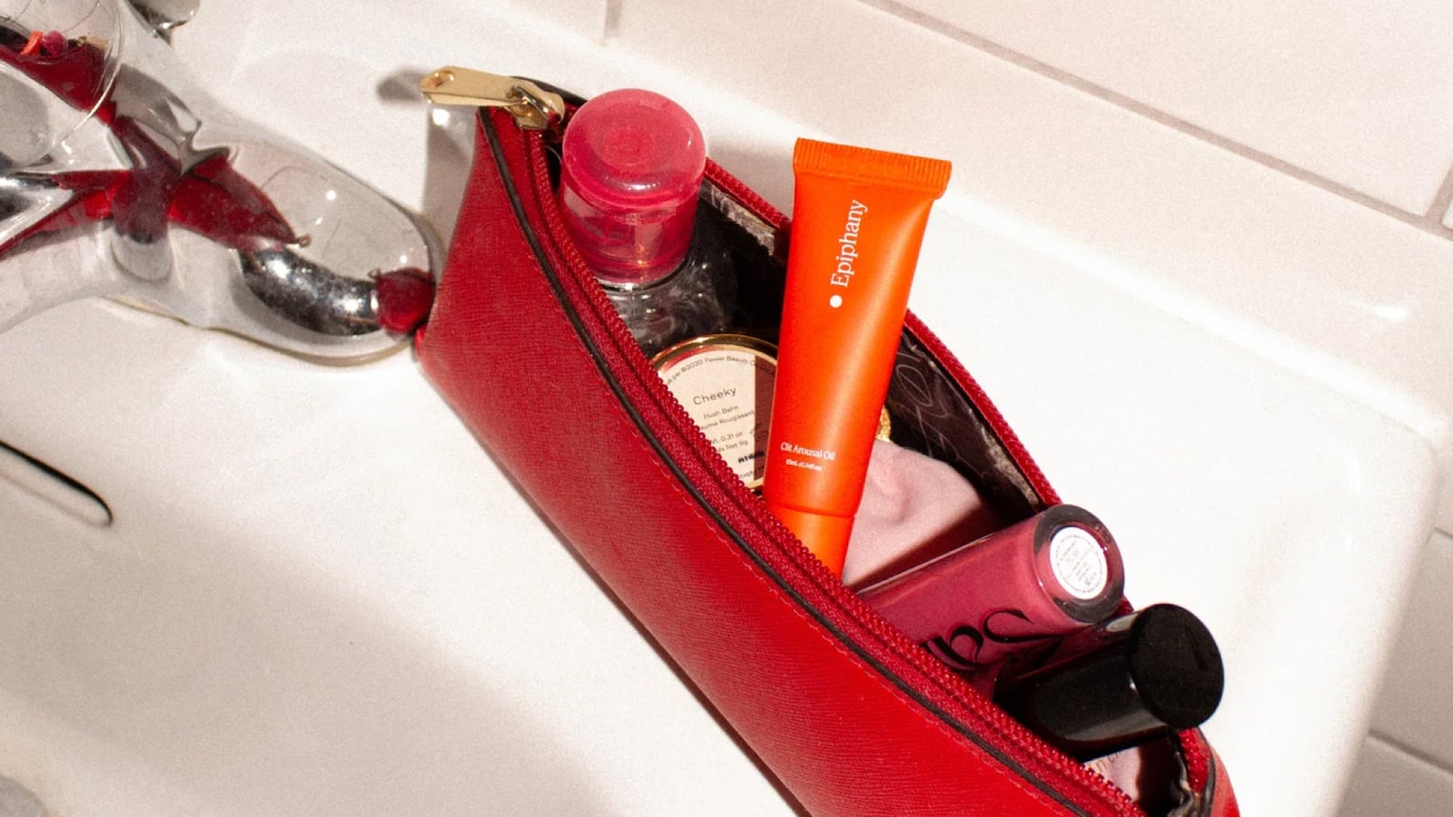 Epiphany Clit Arousal Oil tube tucked inside a red leather makeup bag alongside lip gloss, blush, and other beauty essentials, resting on a bathroom sink.