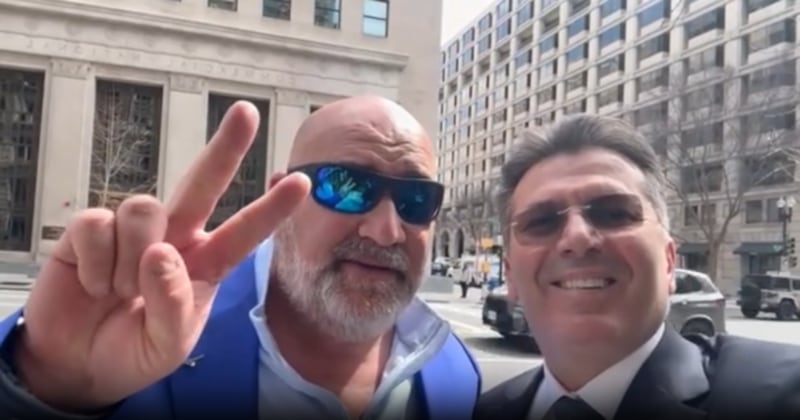 A bald man in sunglasses flashes a V for victory beside a man to his right who has hair and shaded glasses and is smiling. They are in front of office buildings.