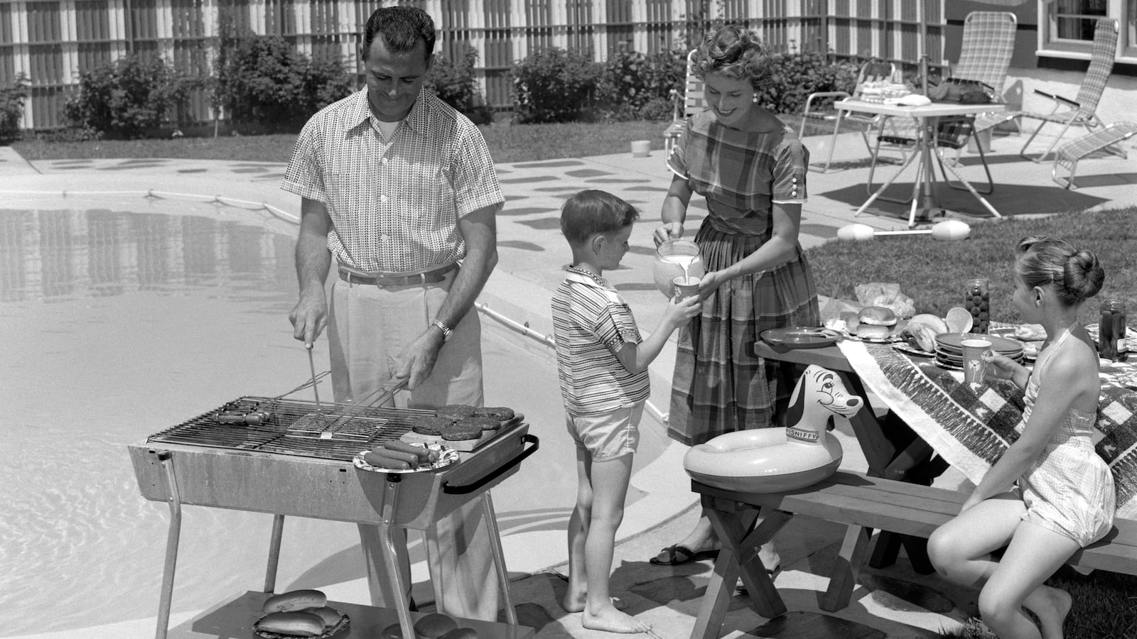 A 1950s family scene of a father grilling by a pool with his wife, son and daughter standing close by. In the background is a ranch-style home.