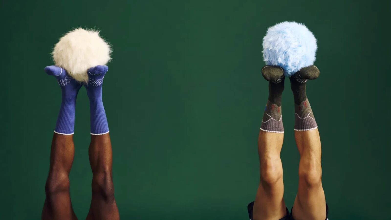 Two pairs of feet, coming in from the bottom of the frame, wearing socks. The one of the right is wearing a blue pair, while the one on the right is wearing a green pair. Both pairs of feet are holding up a fuzzy ball.