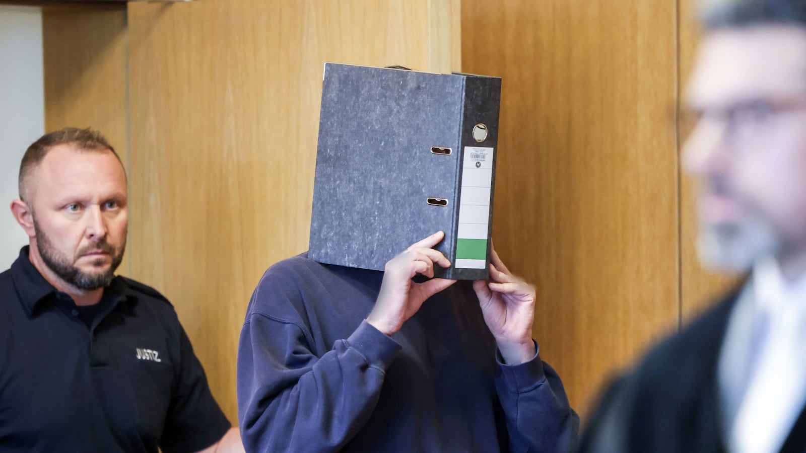 The accused nurse (M) enters the courtroom with a file folder in front of his face.