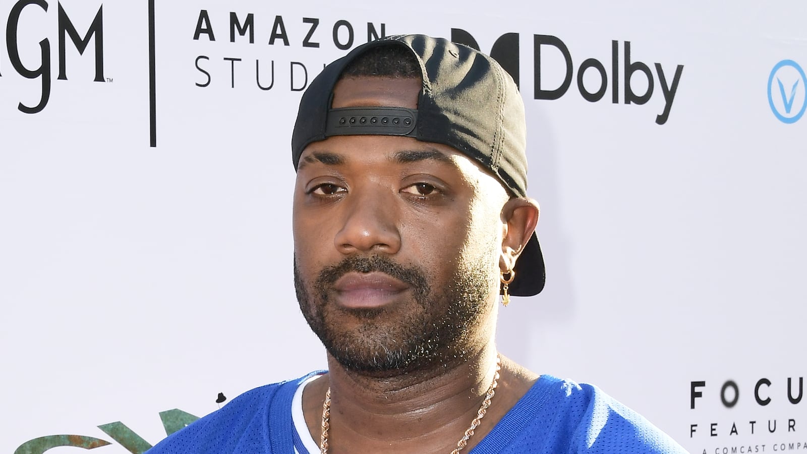 Ray J is in a blue and pink jersey. He is holding up prayer hands. The background has the logos of different companies.