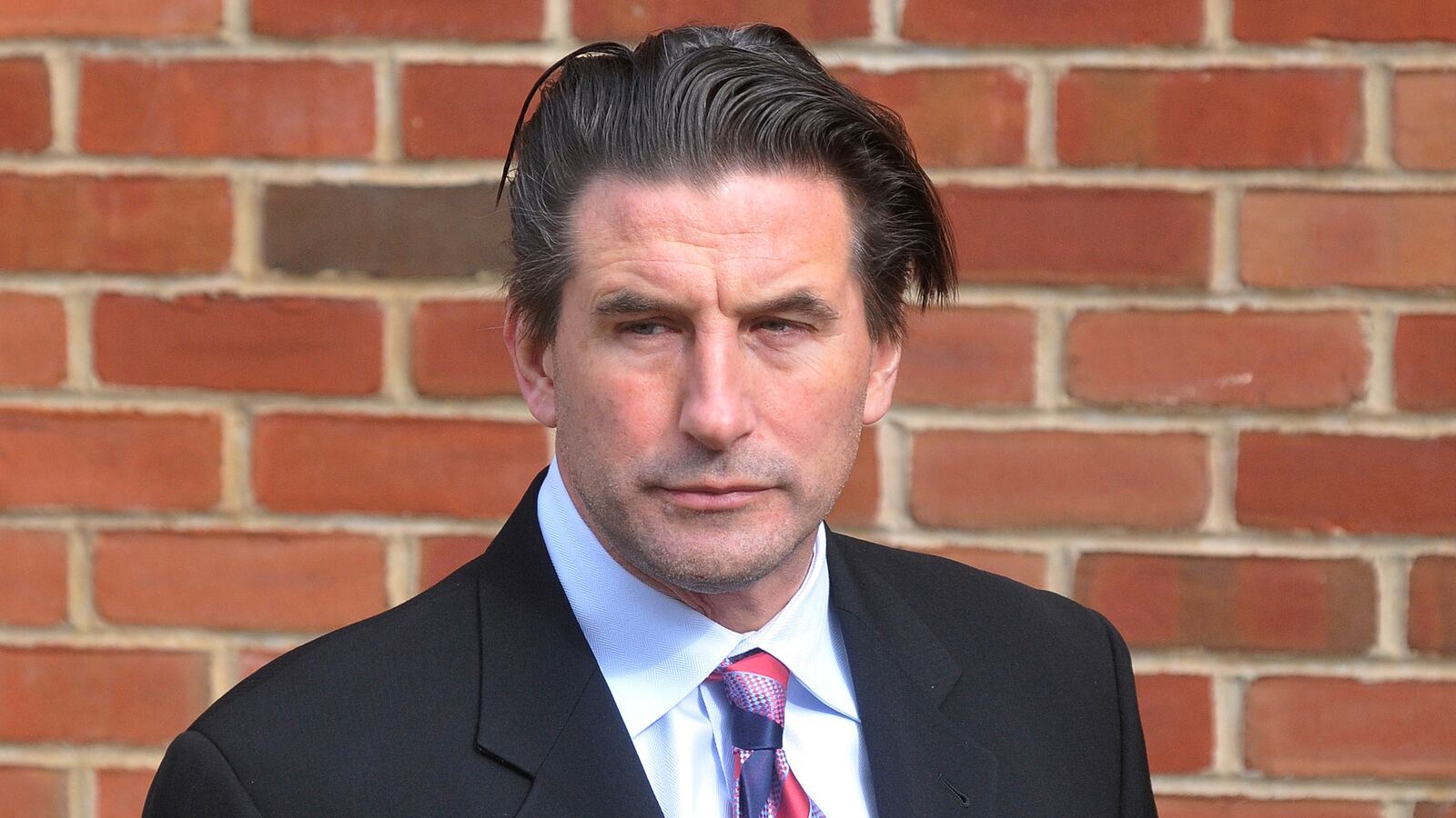 Billy Baldwin wears a suit in front of a brick wall