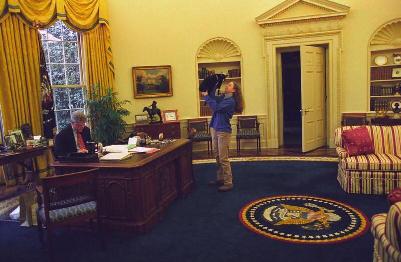 President Bill Clinton, wearing suit and glasses, works at his desk in the Oval Office, while the First Daughter Chelsea Clinton, wearing khaki trousers and a blue sweater, stands facing him with arms stretched upward cradling the First Pet, Socks the Cat, with black fur, white face, and tiny collar, inside the White House, Washington, District of Columbia, December 24, 1994.