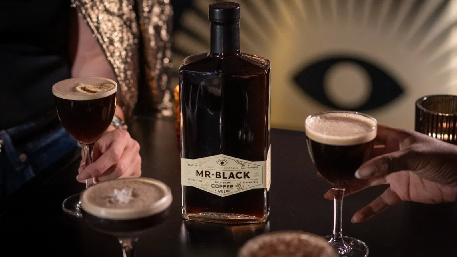 A table with four cocktails and a black bottle of Mr Black Cold Brew Coffee Liqueur on it. Two of the drinks are being held by hands.