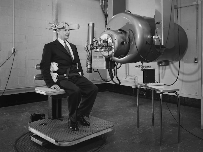 John G. Trump sits next to the compact electrostatic generator he and Dr. Robert J. Van de Graaff developed for use in disease treatment circa 1950. The apparatus, designed to rotate the patient, consists of a platform with a seat and a headrest and a generator mounted from the ceiling and enclosed in a sealed tank.
