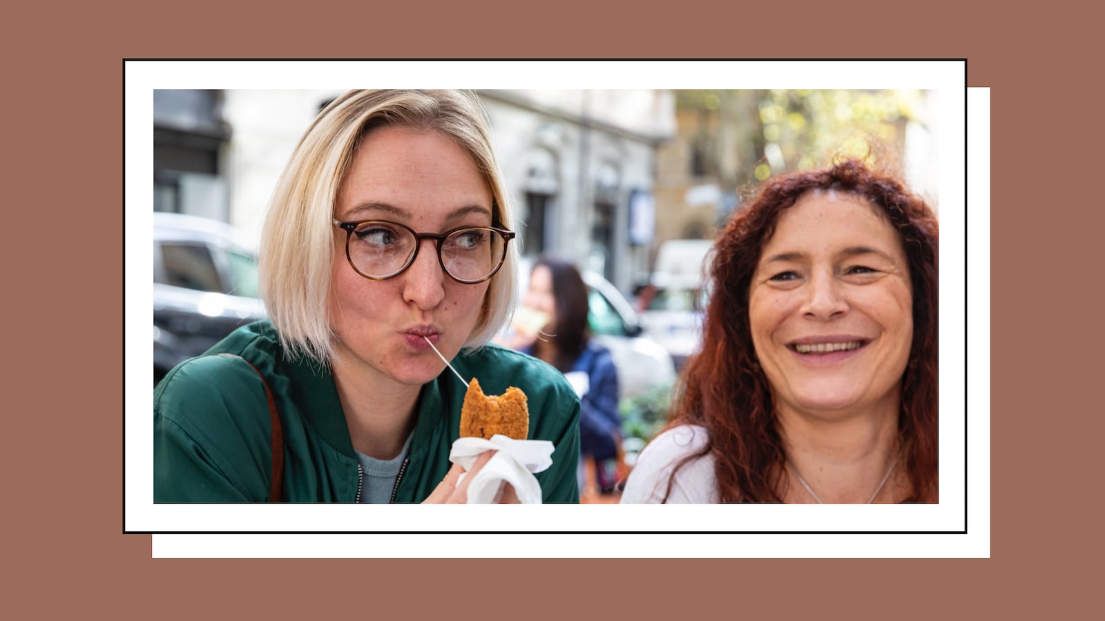 Two women enjoying food together outdoors. One woman with short blonde hair, glasses, and a green jacket holds a fried snack and playfully sips a string of melted cheese. The other woman with long, curly red hair smiles widely. They appear to be sharing a lighthearted moment on a sunny day.