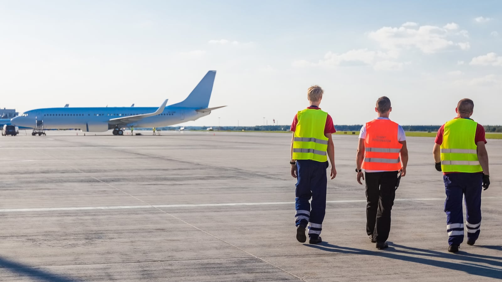 Rear view of three men walking on the airport runway with airplane in the background. Wide angle view.