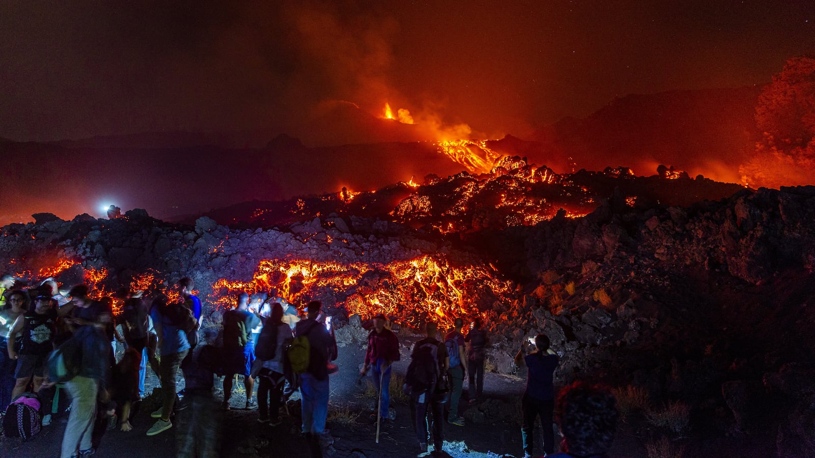 CATANIA, ITALY - AUGUST 14: Mount Etna's eruptive activity continues, with active lava flows fed by vents located at 2,750 and 1,900 meters of altitude, alongside ongoing ash emissions from the Voragine summit crater in Catania, Sicily, Italy on August 14, 2026. The spectacular glowing lava streams have attracted an exceptional influx of hikers, tourists, and curious onlookers eager to witness the volcanic phenomenon at night. In response to this dangerous mass exodus towards the active lava fronts, local authorities have implemented strict roadblocks, parking bans along key mountain routes like the Mareneve road, and restricted access to the affected areas to ensure public safety. Meanwhile, the continuous emission and fallout of volcanic ash have severely impacted operations at Catania International Airport, leading to a total block described as the most severe emergency in the last twenty years, with flights temporarily suspended until tomorrow. (Photo by Salvatore Allegra/Anadolu via Getty Images)