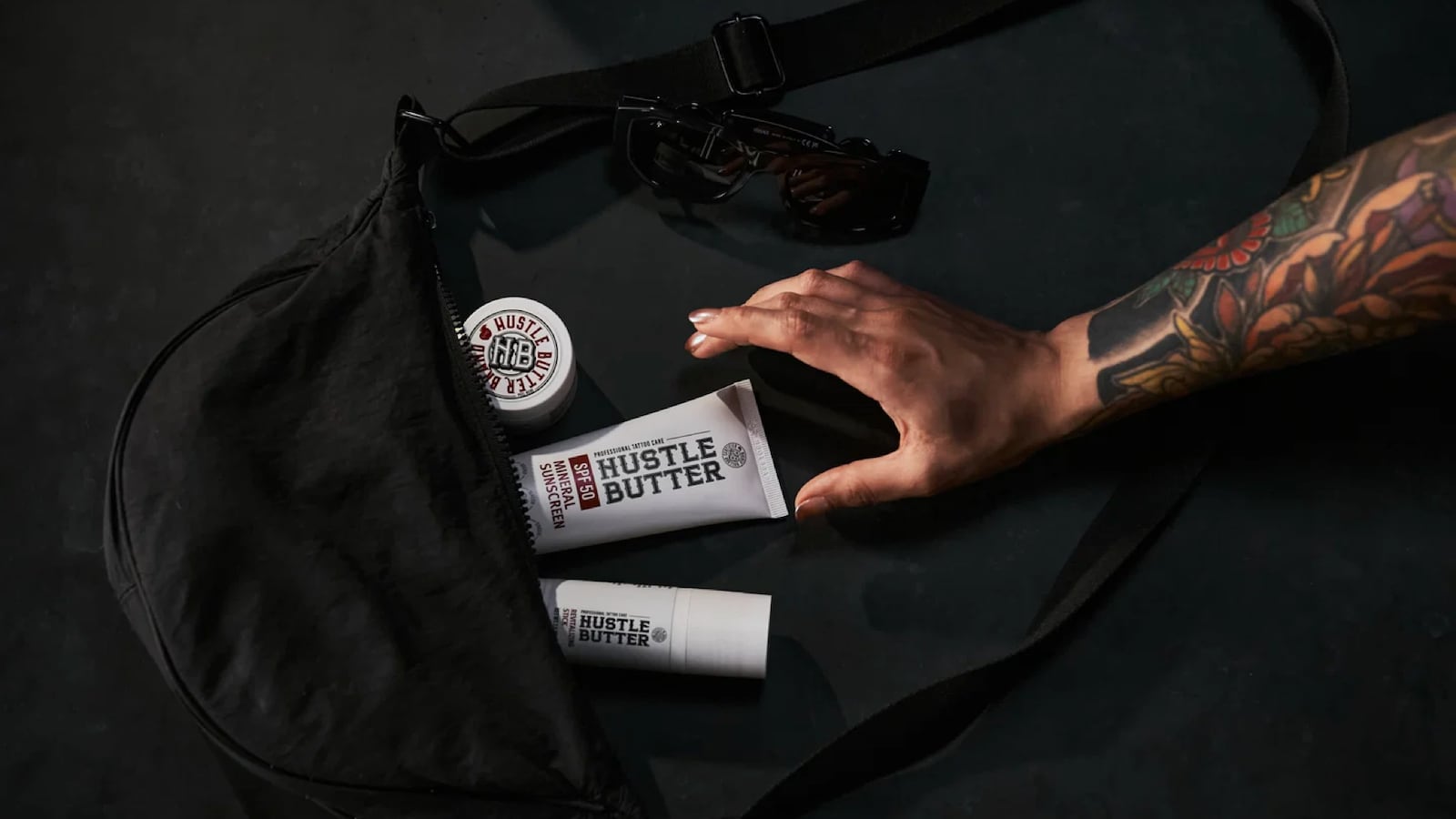 Tattooed arm reaching into a black gym bag containing Hustle Butter Mineral Sunscreen tube, travel-sized stick, and jar, with sunglasses nearby.
