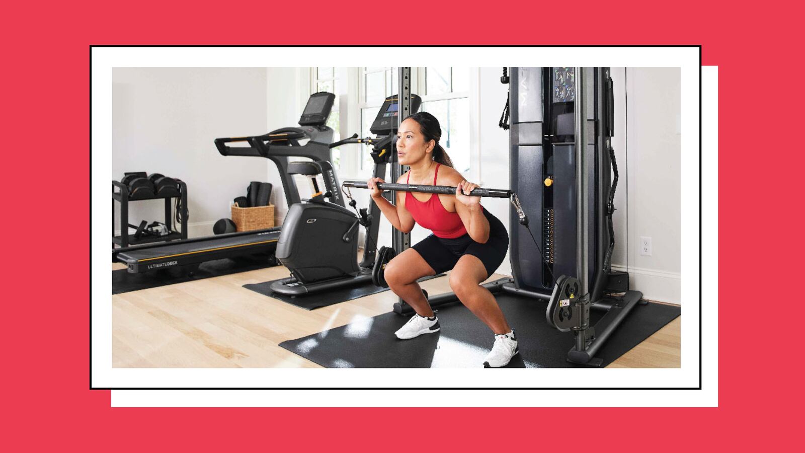 A woman doing squats in a home gym. She is wearing white sneakers, black shorts, and a red top. In the background is a treadmill and elliptical.