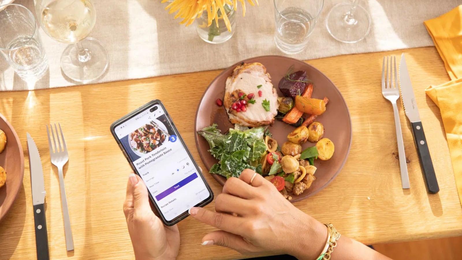 Person using the Weight Watchers app on a smartphone while eating a balanced meal with roasted chicken, vegetables, and salad at a dining table.