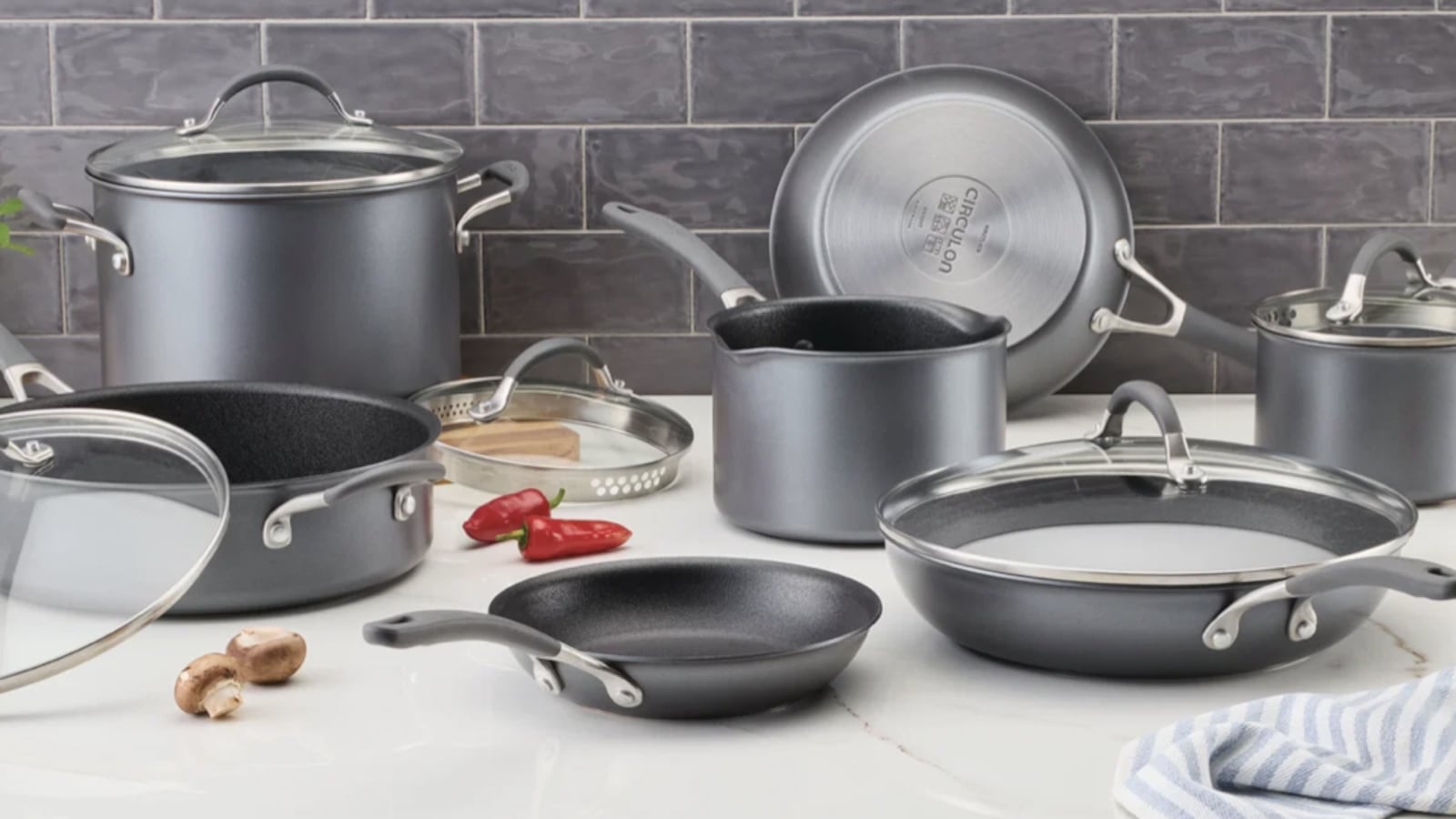Circulon ScratchDefense nonstick cookware set arranged on a white marble countertop against a gray subway tile backsplash, with a straining lid, mushrooms, red chili peppers, and a striped kitchen towel.