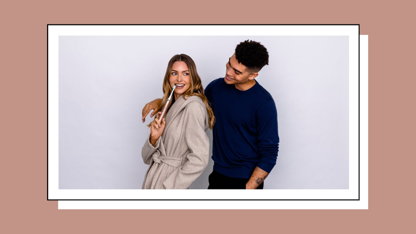 A smiling woman in a beige robe holds an AquaSonic electric toothbrush while a man in a navy sweater stands behind her with his arm around her shoulder, both posed against a light gray background.