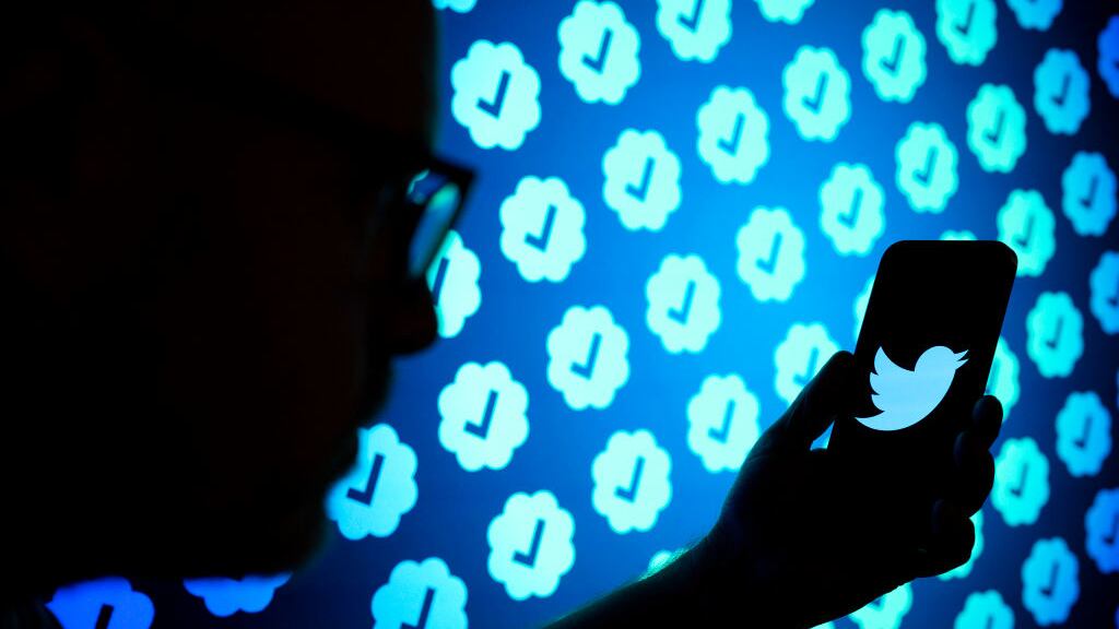 A photo illustrating showing a shadowy figure holding a phone with a Twitter blue bird logo. In the background, there is repetitive imagery of Twitter’s verified badges. Elon Musk recently announced a temporary limit on the number of posts users can read.