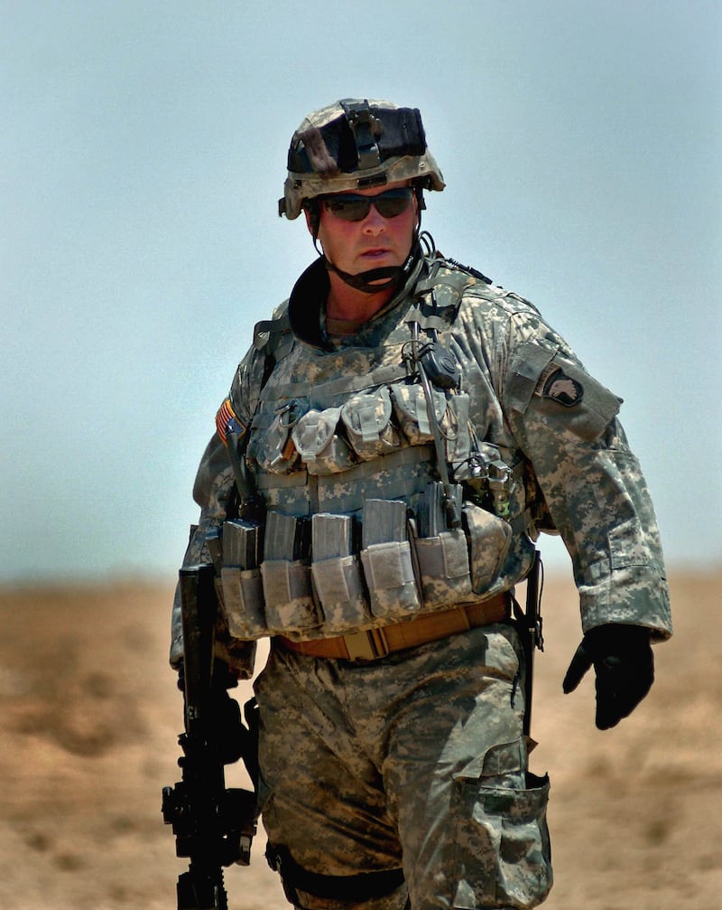 A man in green uniform and ballistic helmet holding a military issue rifle in a desert.