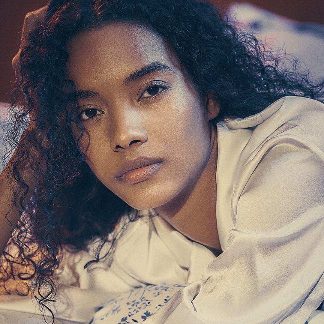 Close-up of a woman with natural curly hair lying on a bed, wearing a cream-colored satin blouse, gazing calmly into the camera with soft evening light illuminating her face.