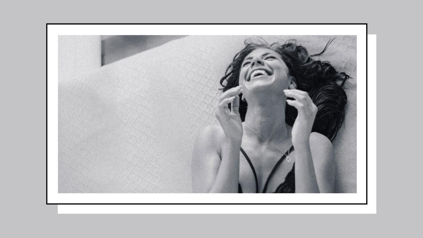 A black and white photo of a woman wearing lingerie while lying on top of a bed. She is laughing.