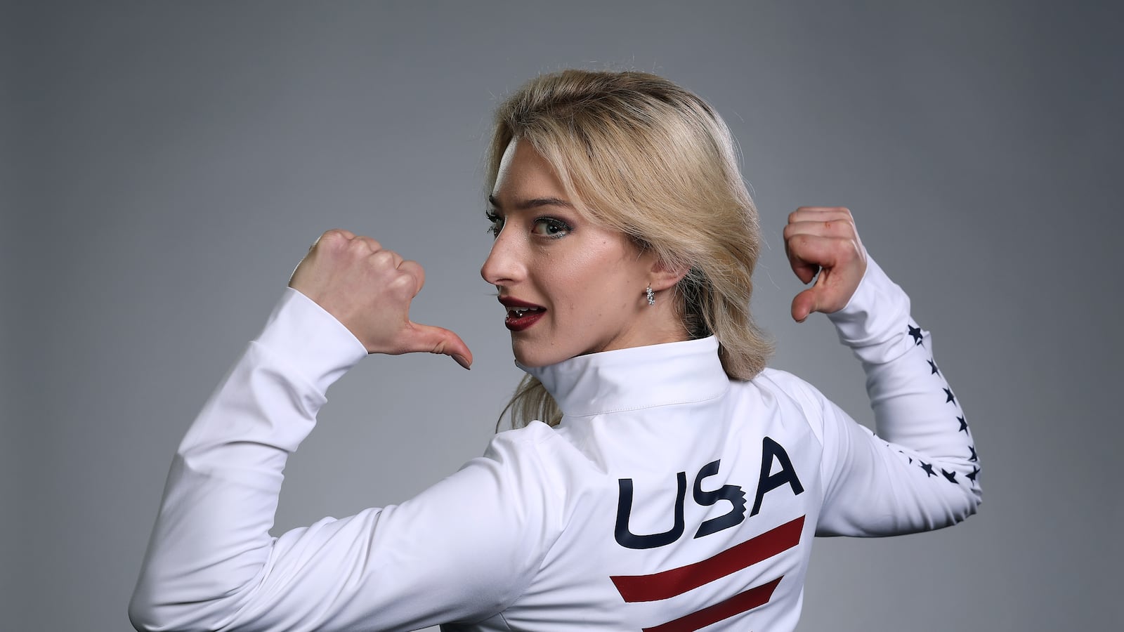 A blonde woman faces away from the camera pointing to the 'USA' logo on the back of her jacket.
