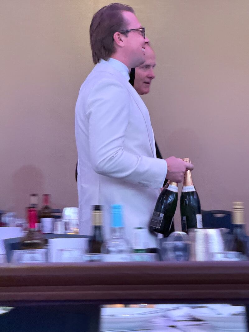 Unidentified gentleman in white tuxedo carries 2 bottles of champagne.