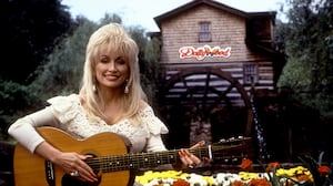 Dolly Parton poses with her guitar in front of Dollywood sign.