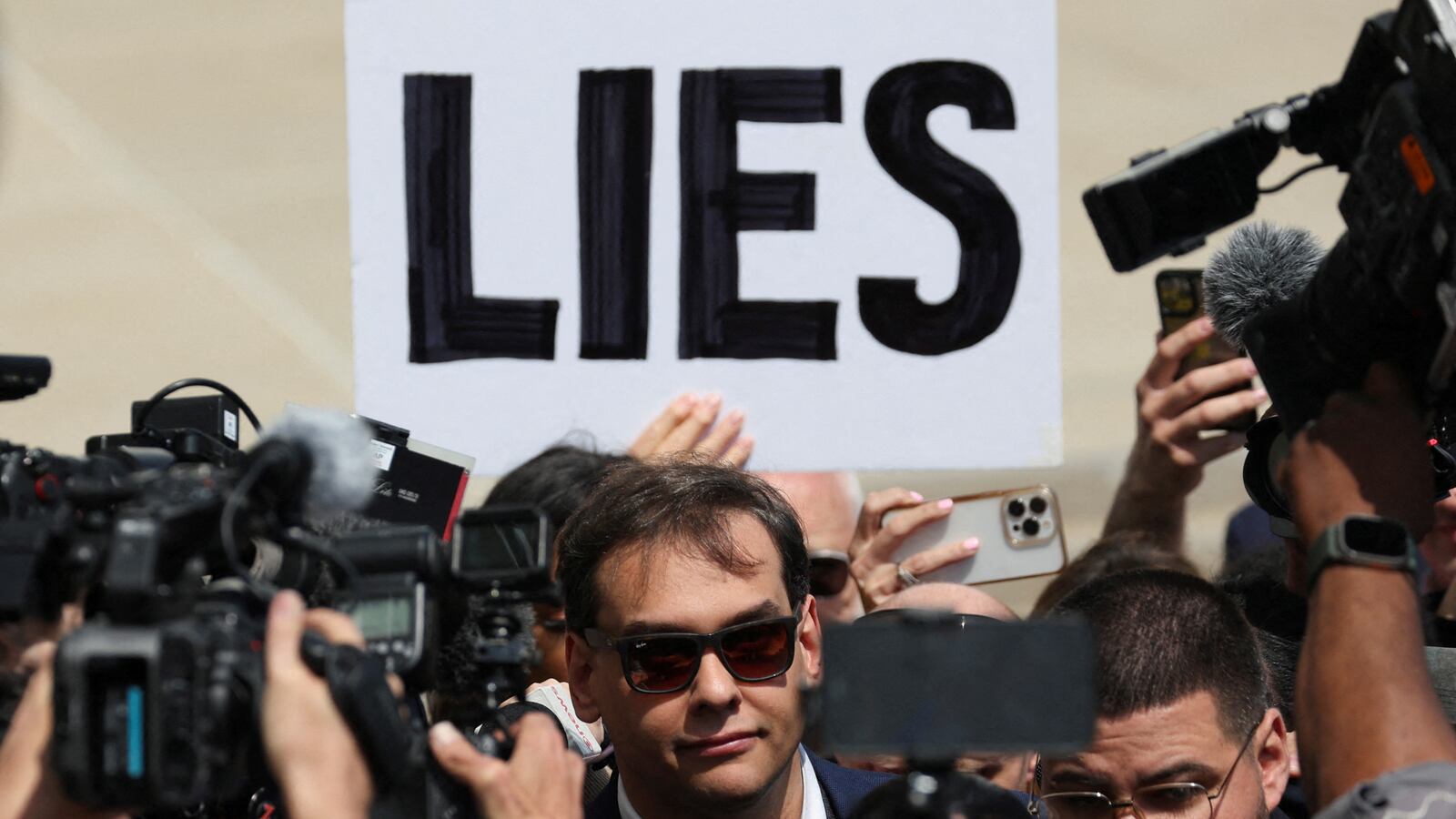 A picture of Rep. George Santos leaving court as a protester holds up a sign reading, “LIES”