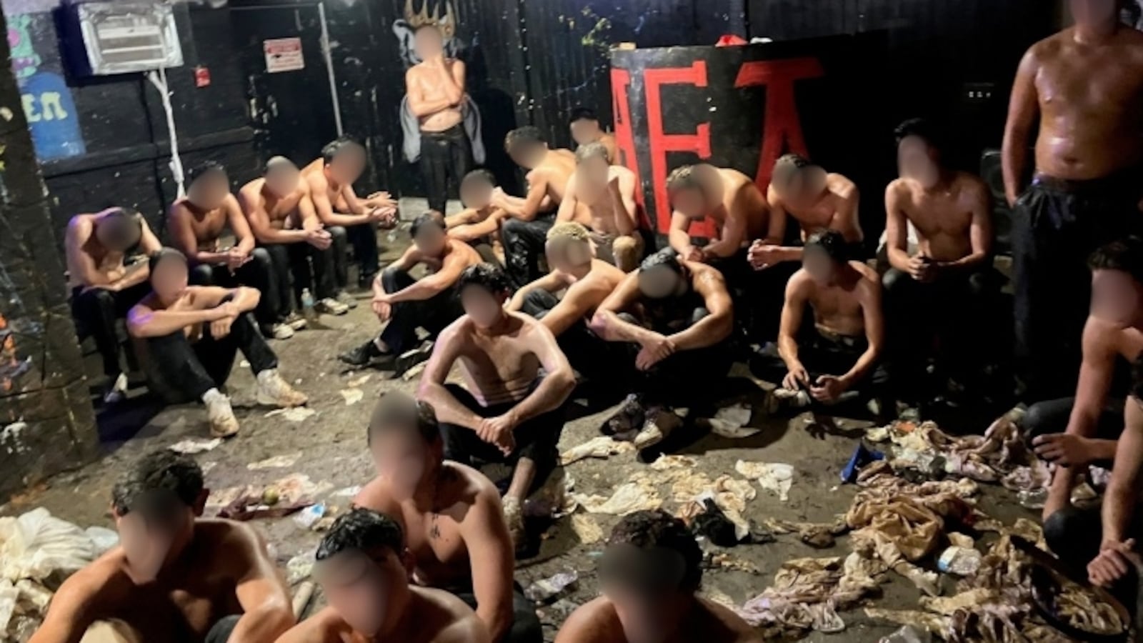 A group of young men without their shirts, faced blurred, sitting in a dirty room.