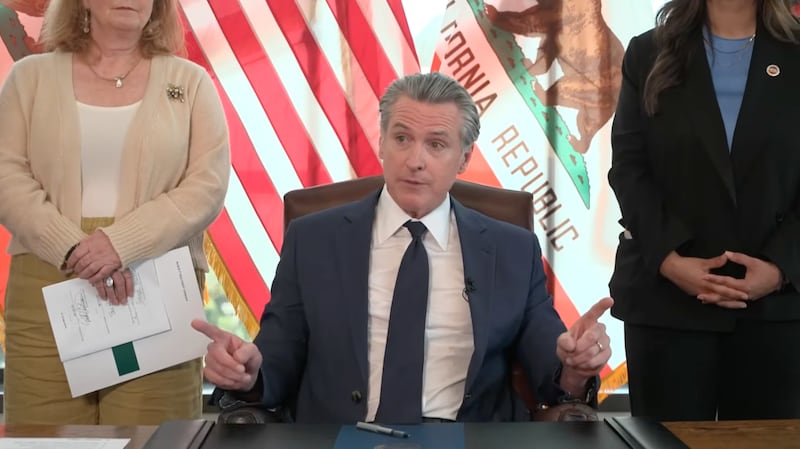 California Governor Gavin Newsom sits at a desk in front of the U.S. and California flags.