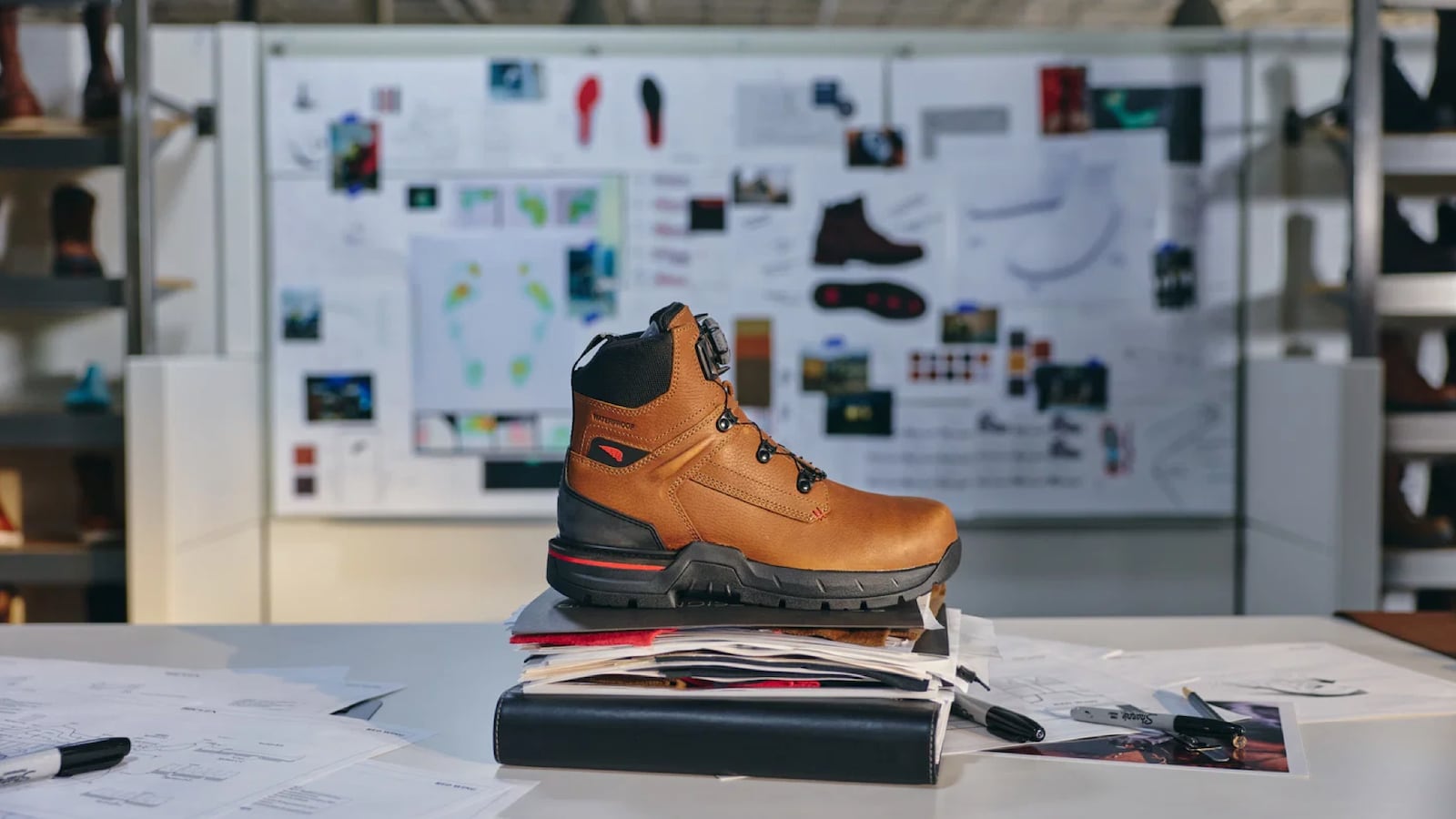A Red Wing IronFlex work boot displayed on a stack of design documents in a footwear studio, with sketches, sole diagrams, and color swatches visible on a mood board in the background.