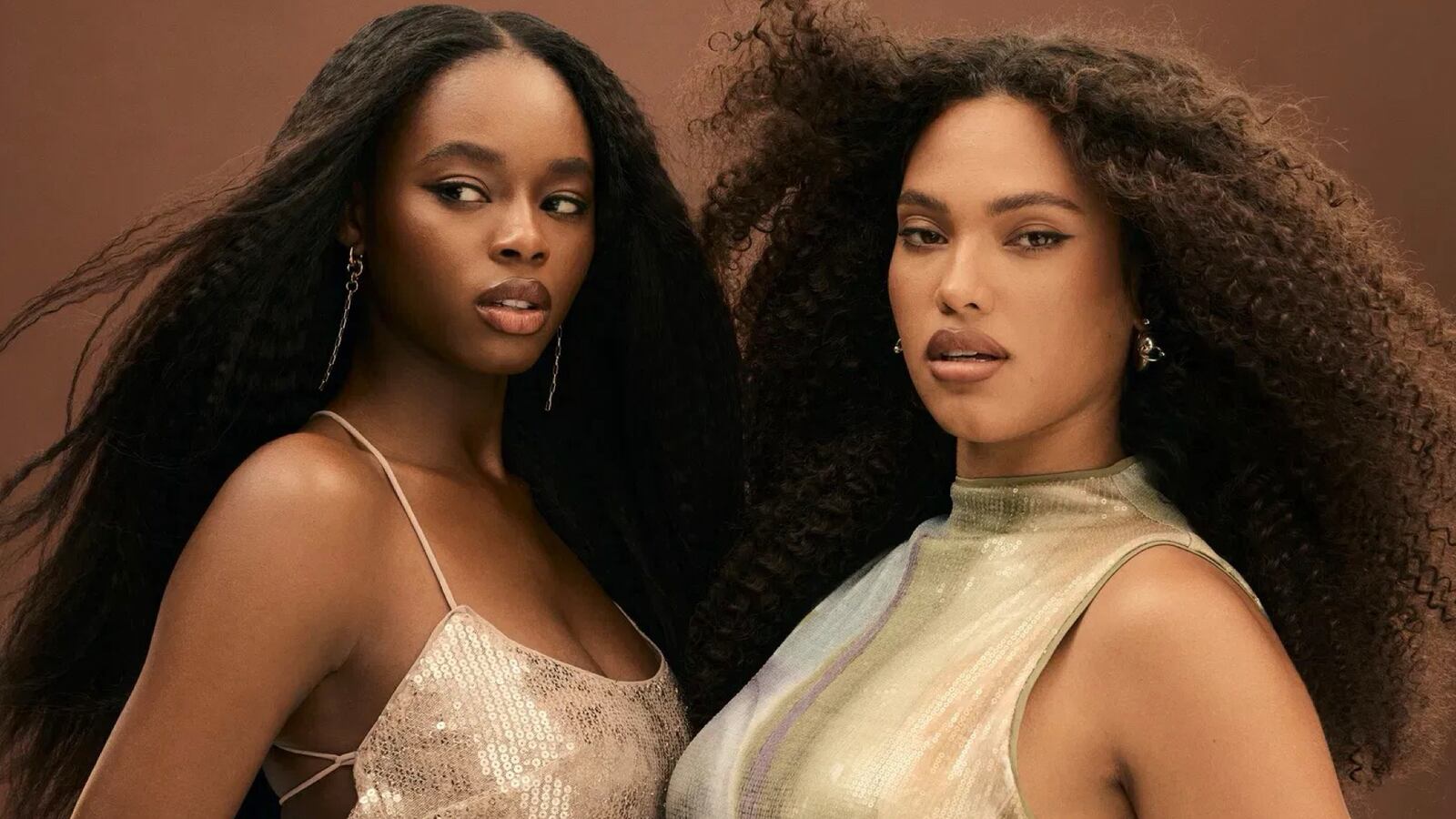 Two Black models in gold dresses smolder. The woman to the left glances to the side, wearing Bellami extensions in a blowout texture. The woman on the right looks at the camera with her curly hair extensions blowing away from her face.