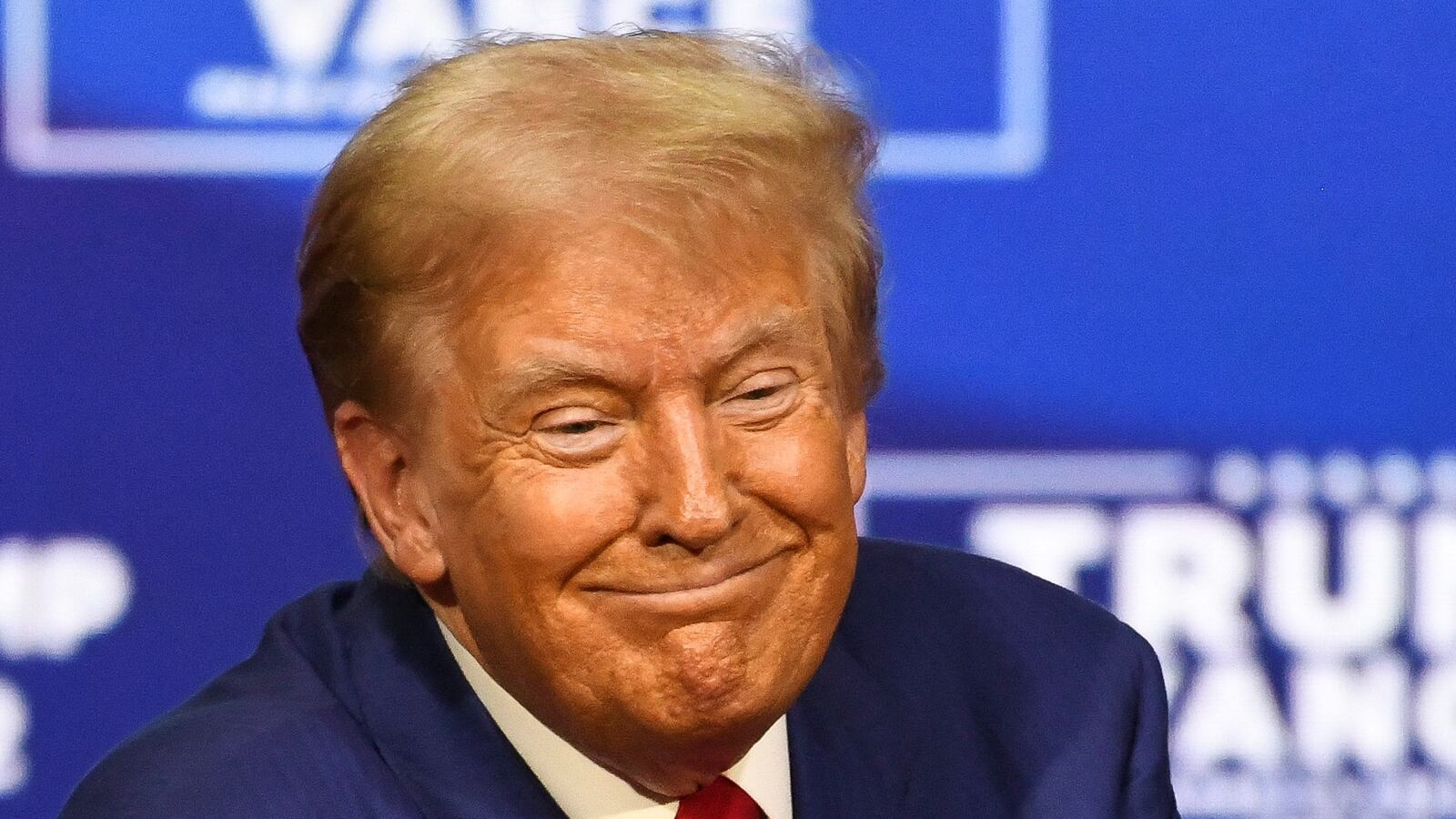 Donald Trump smiles in front of a blue background on stage.