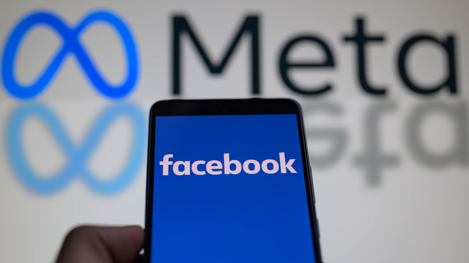 A smart phone is displaying Facebook with the Meta icon visible in the background in this photo illustration.